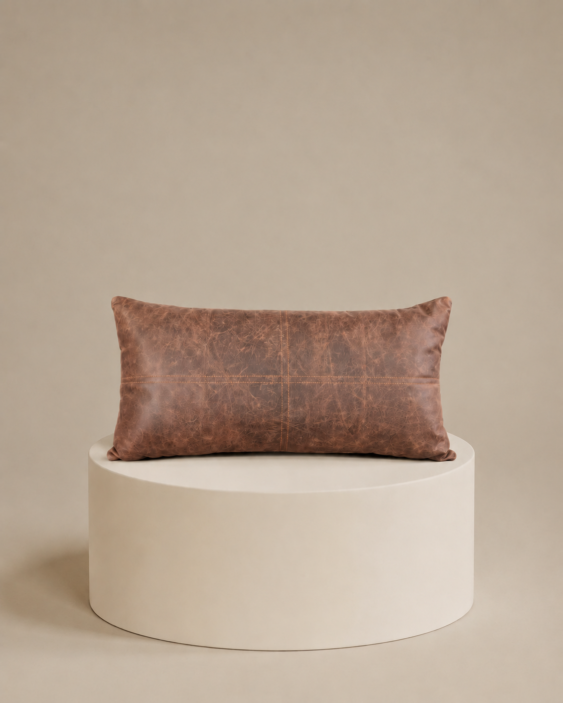 Almohadón de cuero rectangular en color marrón, de 60 × 30 cm, apoyado sobre un pedestal cilíndrico color crema
